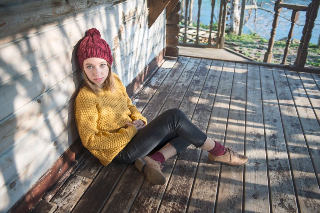 A young woman in a knitted cap and sweater relaxes on a wooden porch in natural light.