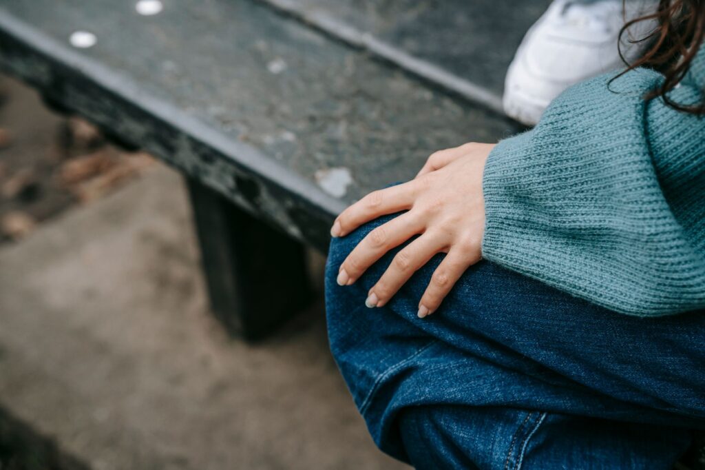 From above of anonymous female in casual outfit sitting on bench with hand on knee while resting on street with unrecognizable person on blurred background