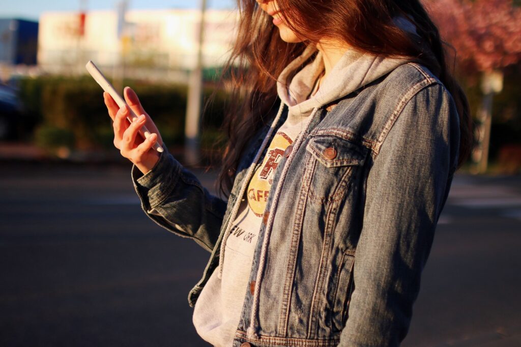 A young woman wears a denim jacket while checking her smartphone outside, showcasing casual fashion.