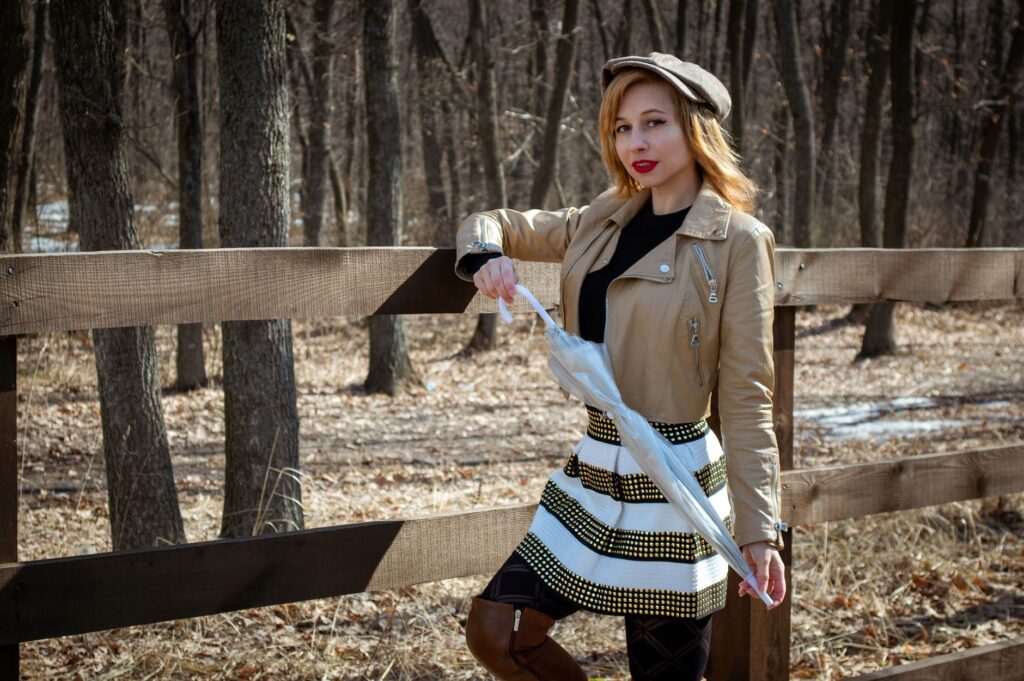 Beautiful young female in stylish clothes and hat standing near wooden fence and looking at camera