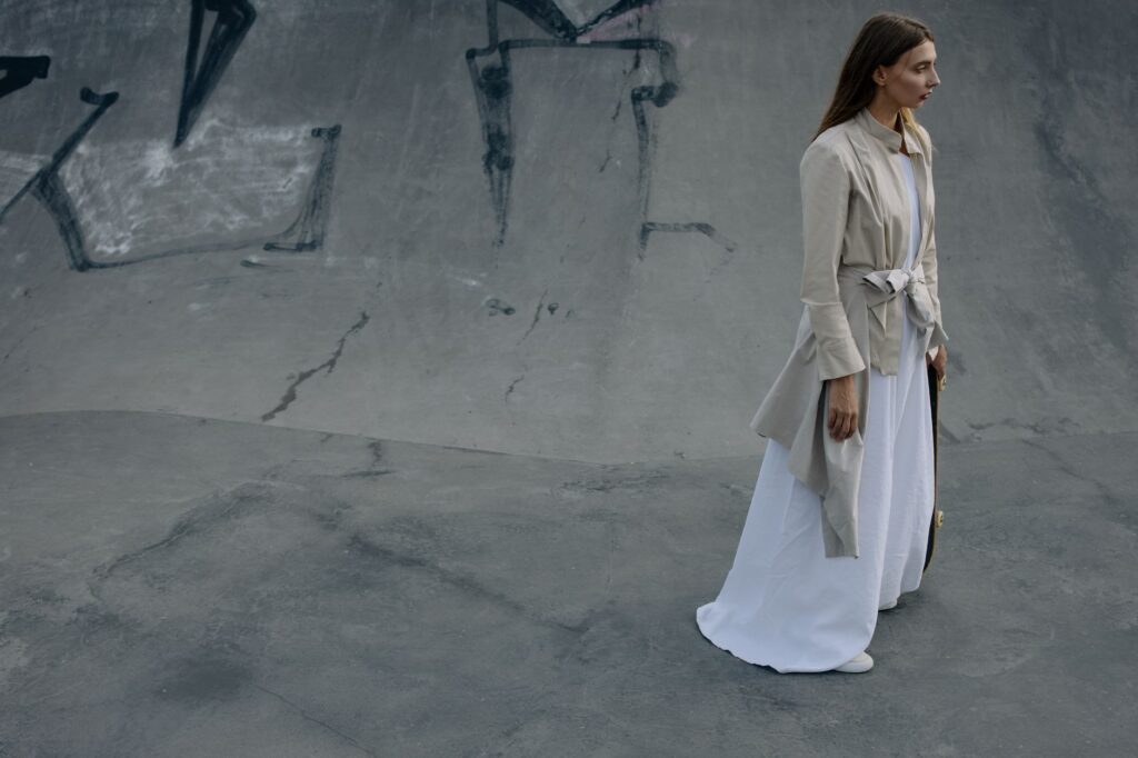 A woman in a stylish beige coat and white dress in an urban concrete setting.