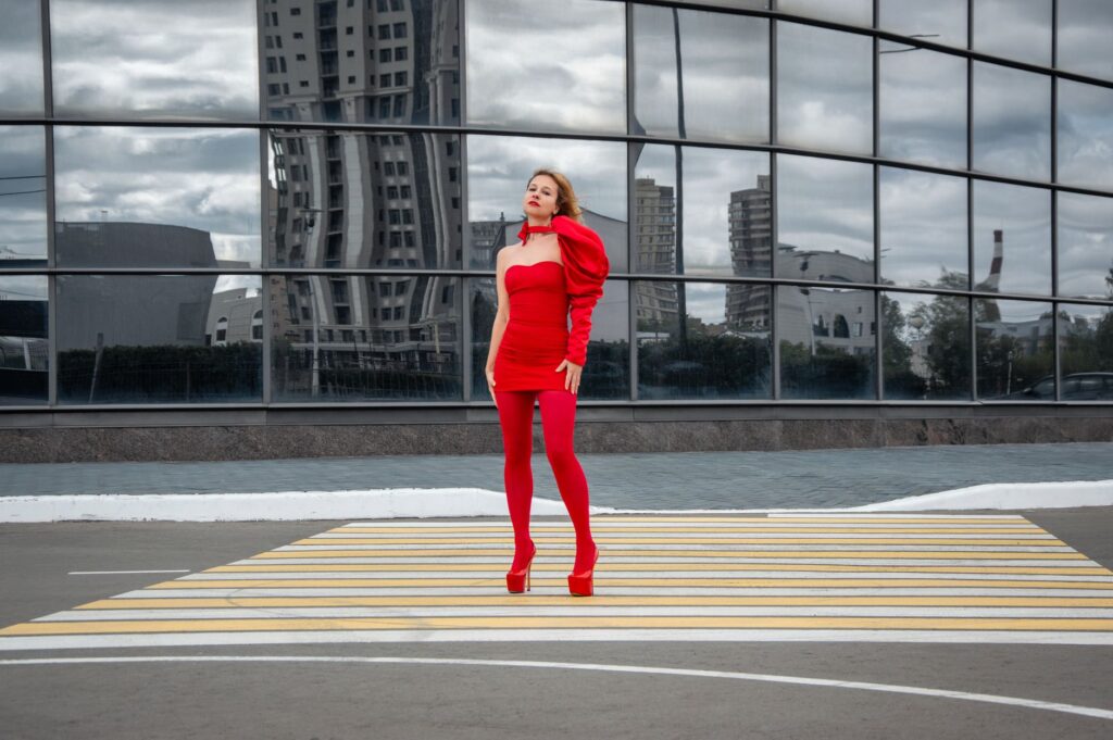 Stylish woman in bold red dress stands confidently in modern urban environment with glass reflections.