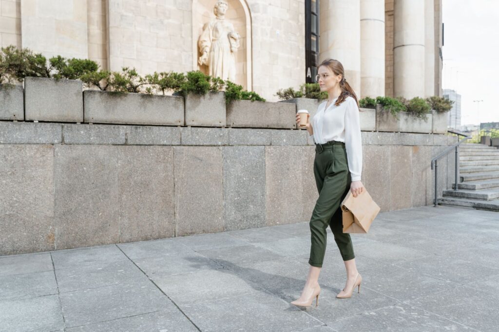 Confident woman in stylish attire walks outdoors holding coffee and documents.
