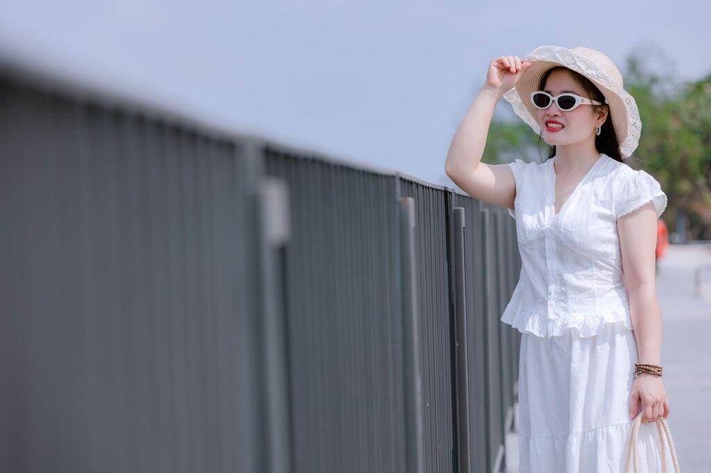 A fashionable woman wearing sunglasses and a hat stands outdoors by a railing on a sunny day.