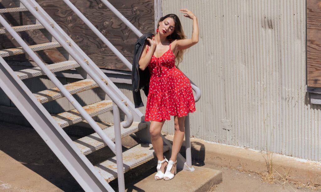 Stylish woman in a red dress poses by industrial staircase under bright sunlight.