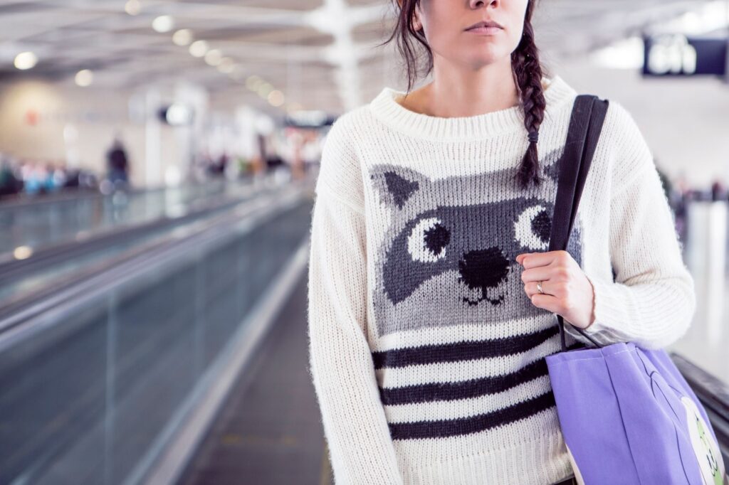 Stylish woman in knit sweater with raccoon design carrying bag in airport.