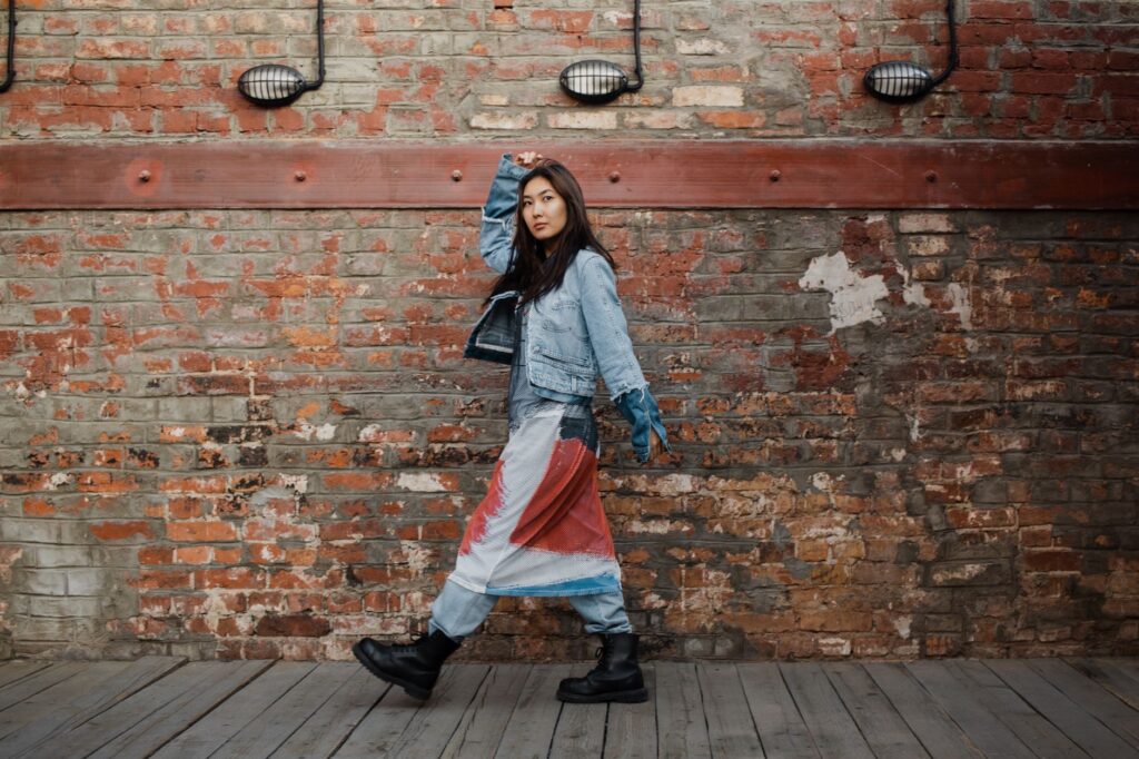 Stylish woman in denim jacket walking past an industrial brick wall.