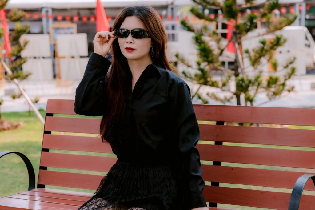 Trendy woman wearing sunglasses sits on a bench outdoors, exuding confidence.