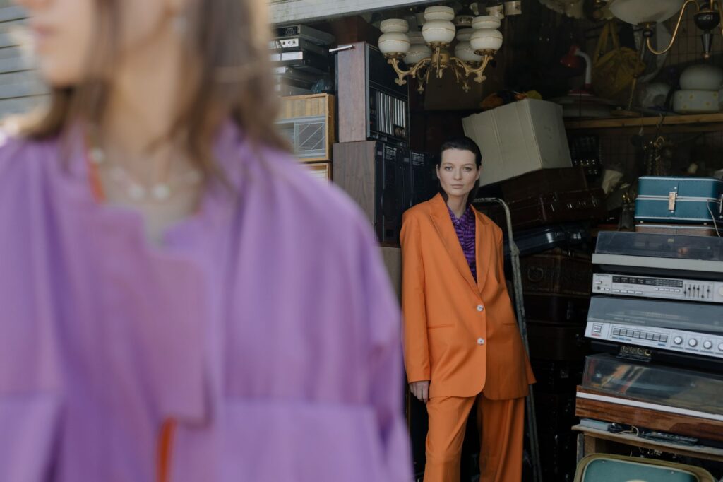 Two women in stylish attire, featuring a striking orange suit, standing amidst vintage electronics and decor.