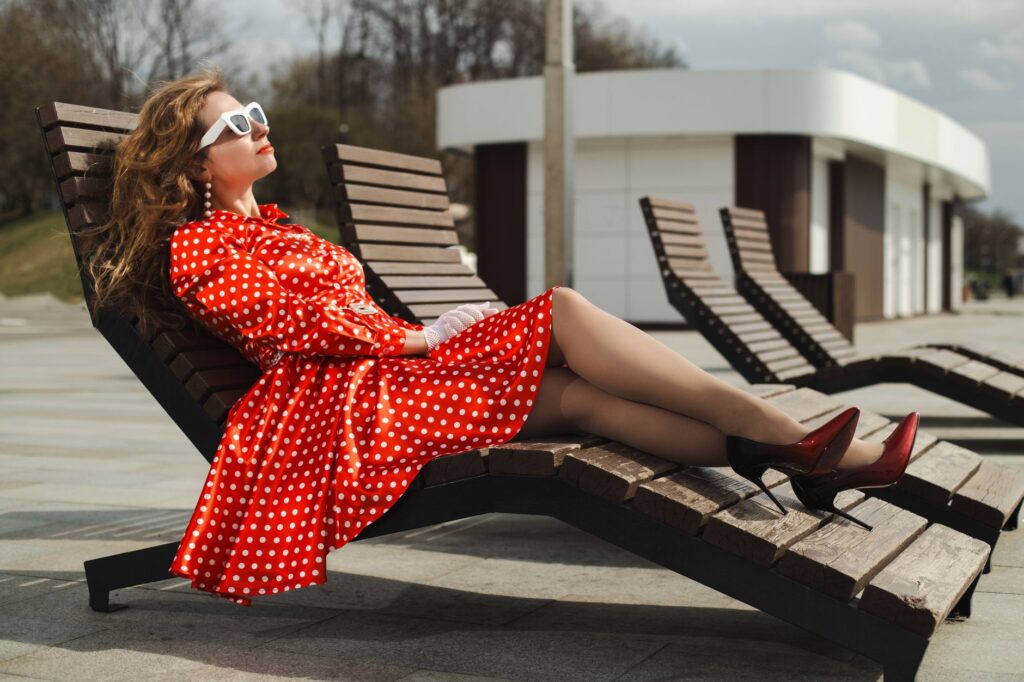 Woman in a red polka dot dress relaxing on a lounge chair outdoors, enjoying a sunny day.