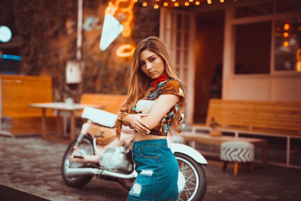 Stylish young woman in denim skirt posing by a motorcycle on an urban street.